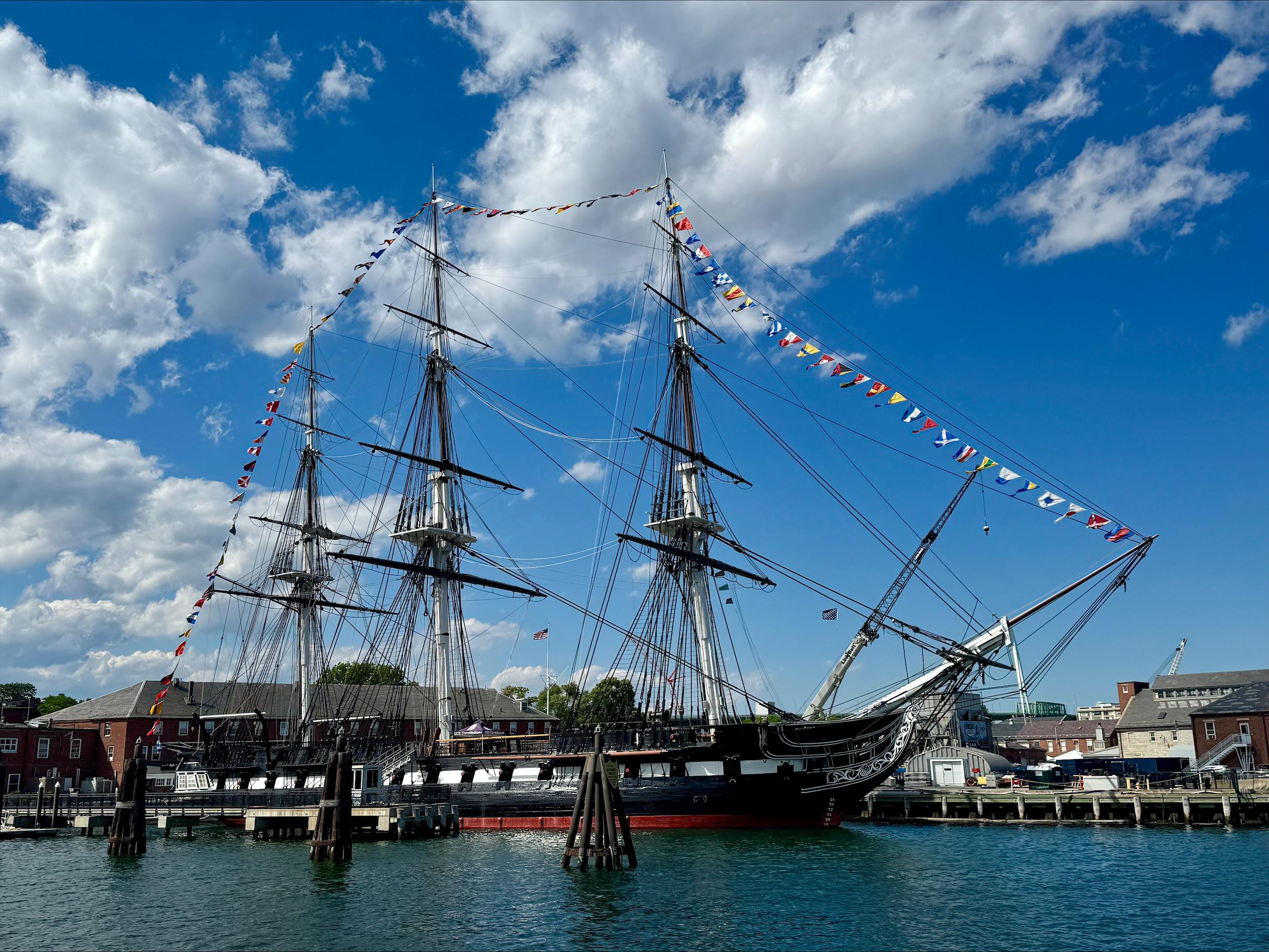 USS Constitution, a three-masted sailing ship, is decorated in flags on a partly-cloudy day.