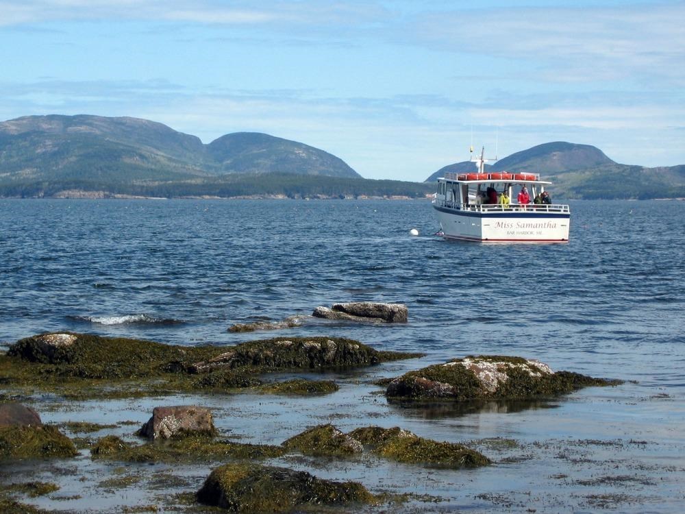 A boat heads out from a rocky shore, with tree-lined mountains in the background