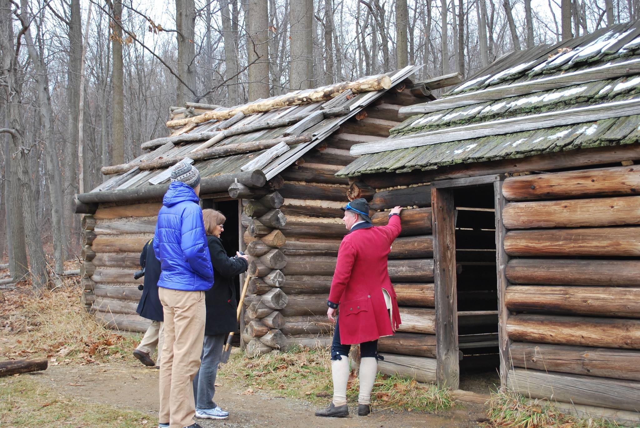 Two men in colonial living history clothing standing in front of log cabins.