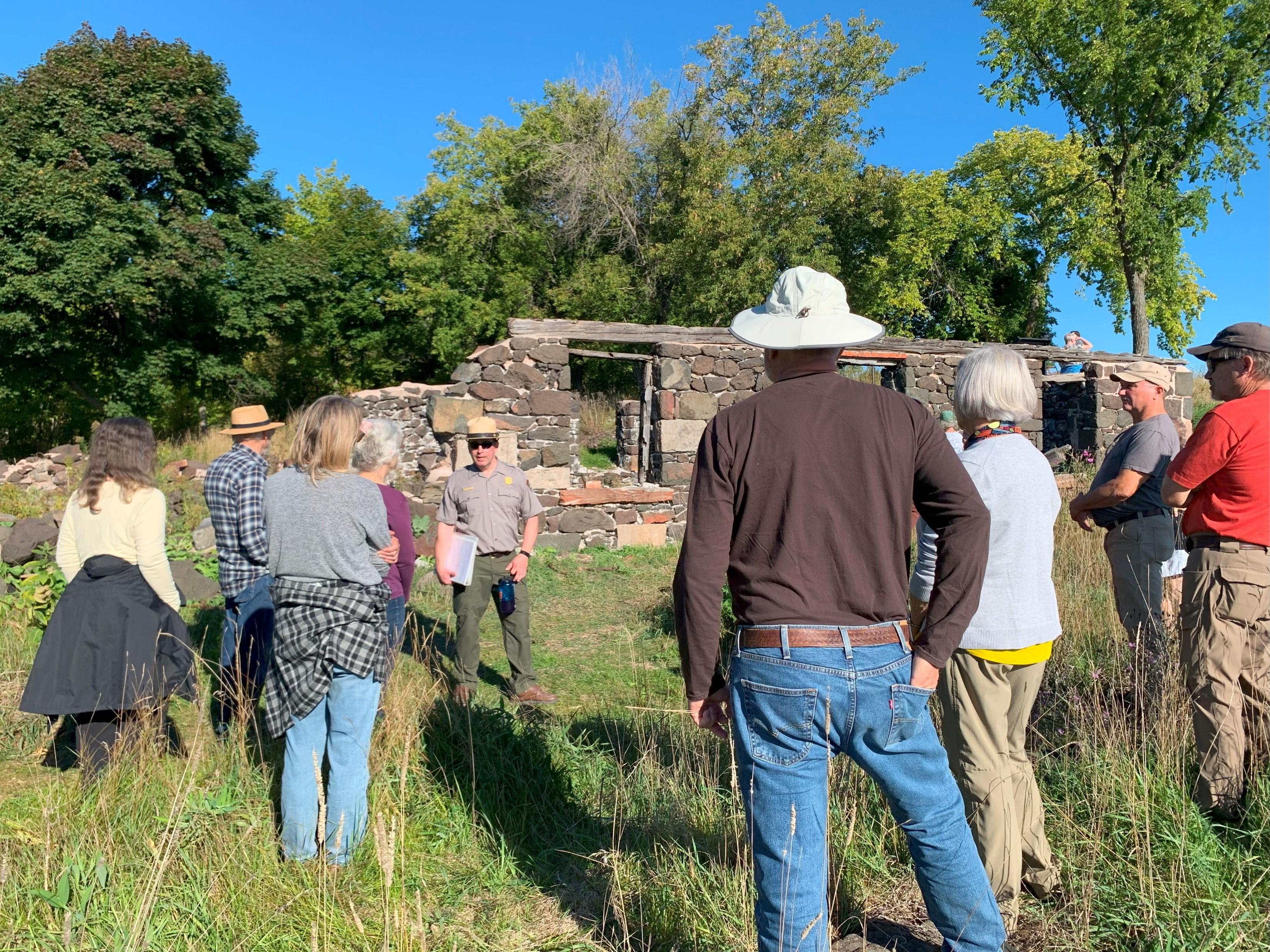 A group stands in a grassy area listing to park ranger. Ruins of a stone building are behind him.