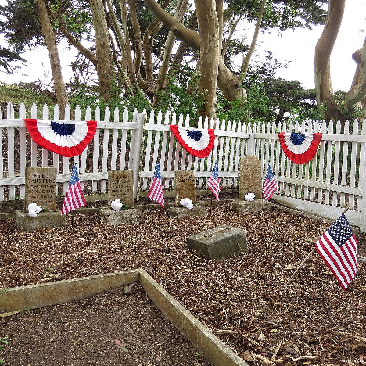 A small cemetery with four headstones surrounded by a white picket fence upon which bunting is hung.