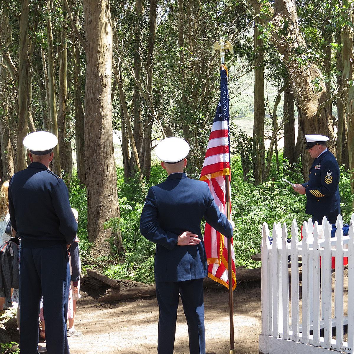 Three U.S. Coast Guard officers in dress uniforms in a wooded area adjacent to a white picket fence.