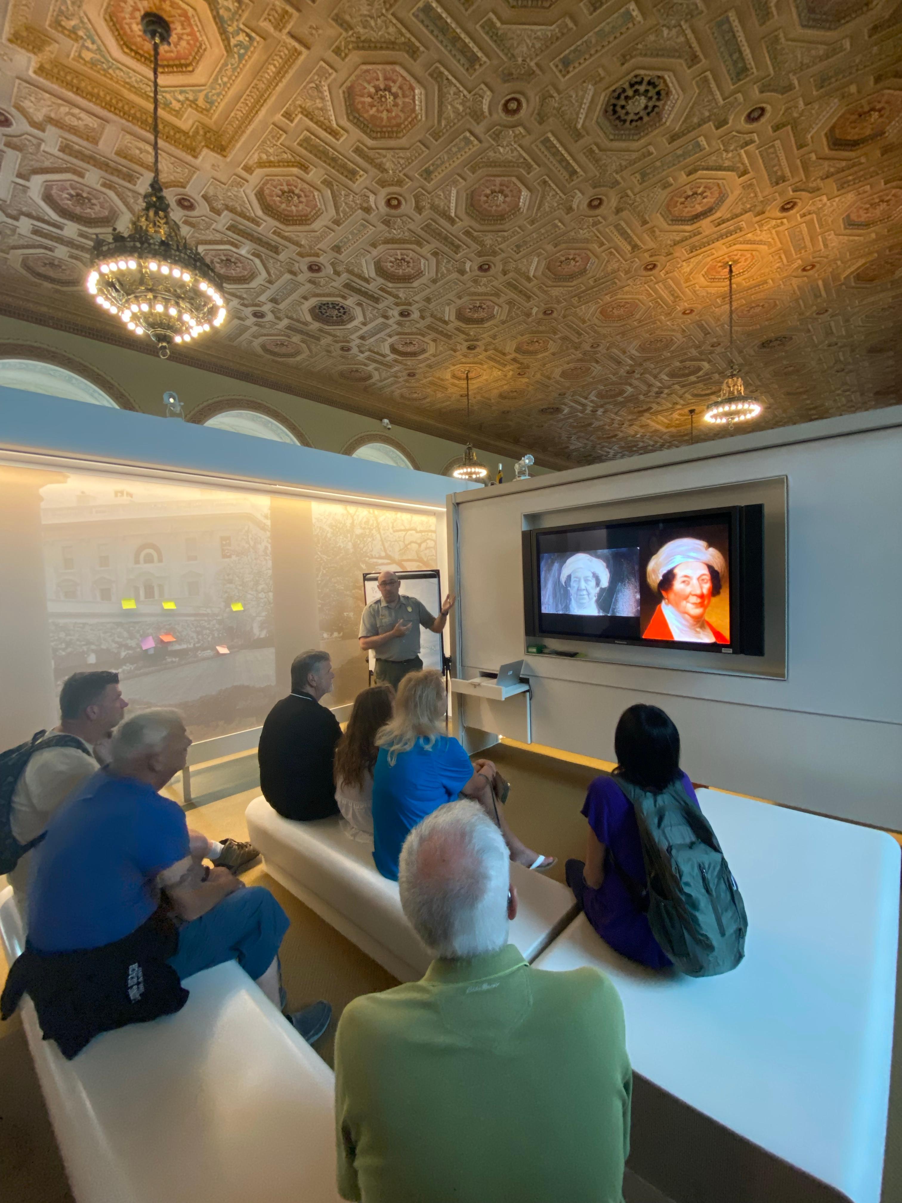 Group of people sits around white benches as a man speaks in front of a televison
