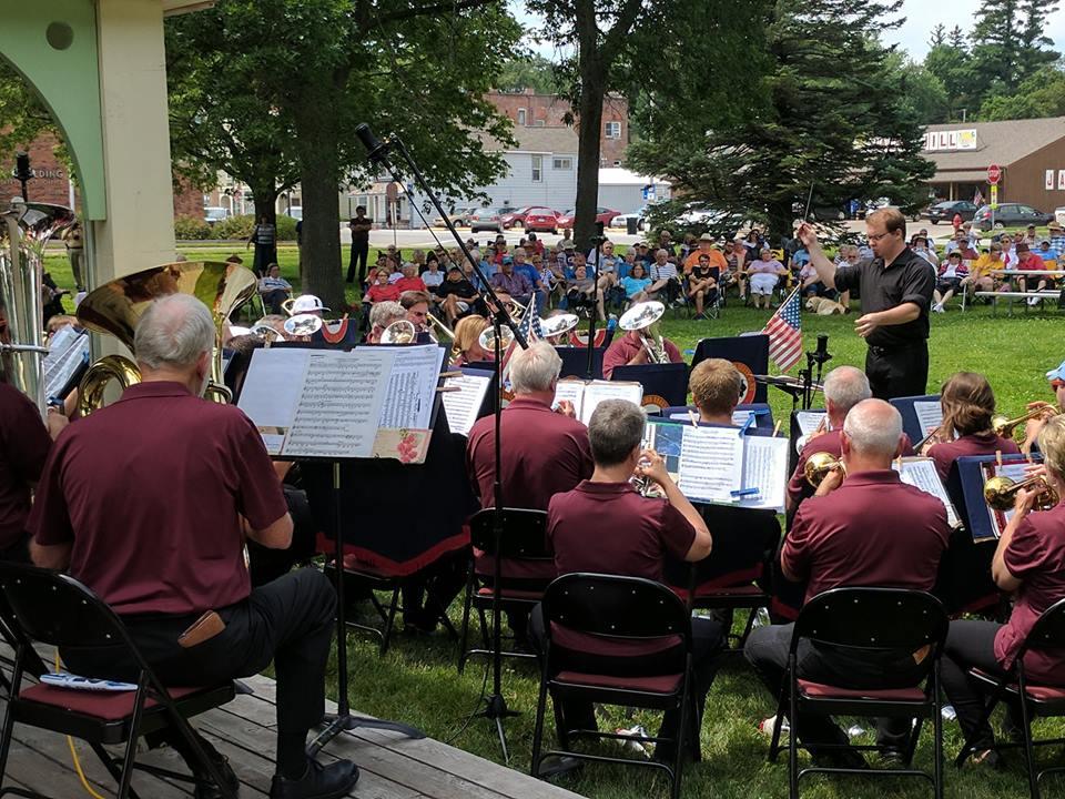 A standing band director wearing black conducts sitting band members playing various horns.