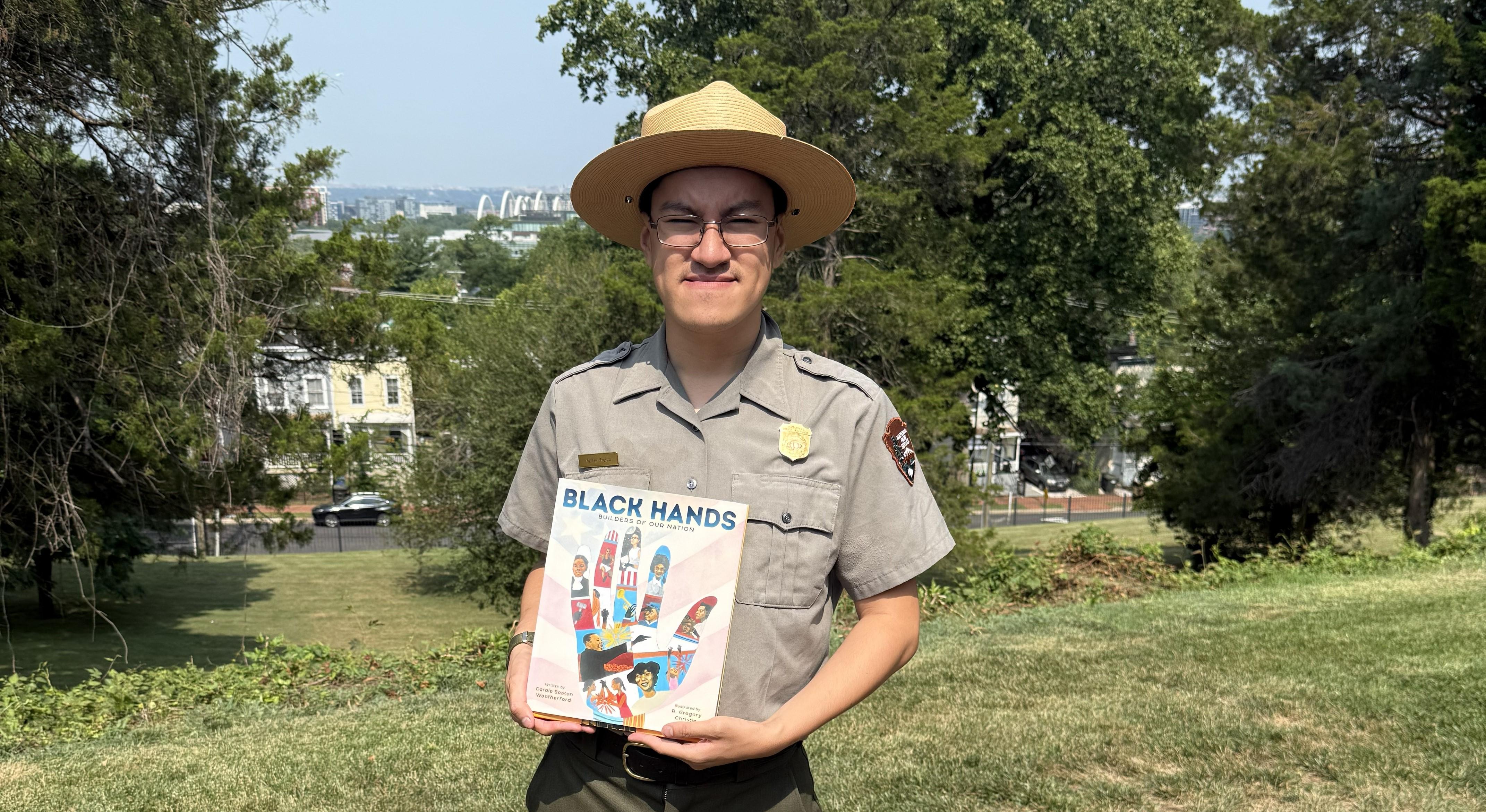 NPS Ranger stands outside holding a book with trees, grass, and blue sky in the background.