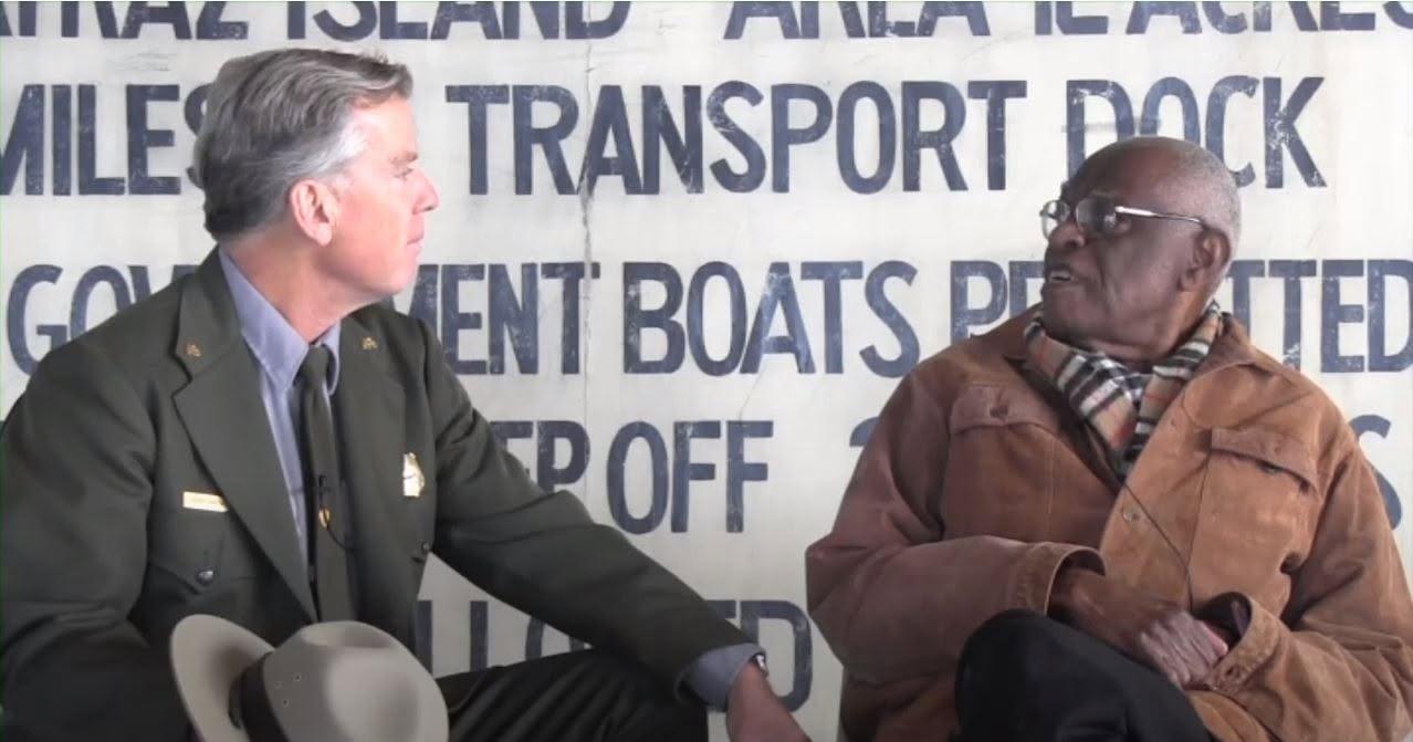 Elderly Black man sits, speaking to a park ranger in green uniform.
