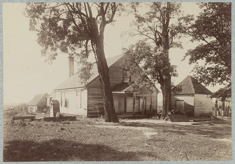 1860s Black and white photo of 2-bay wood frame building with smaller outbuildings and trees nearby