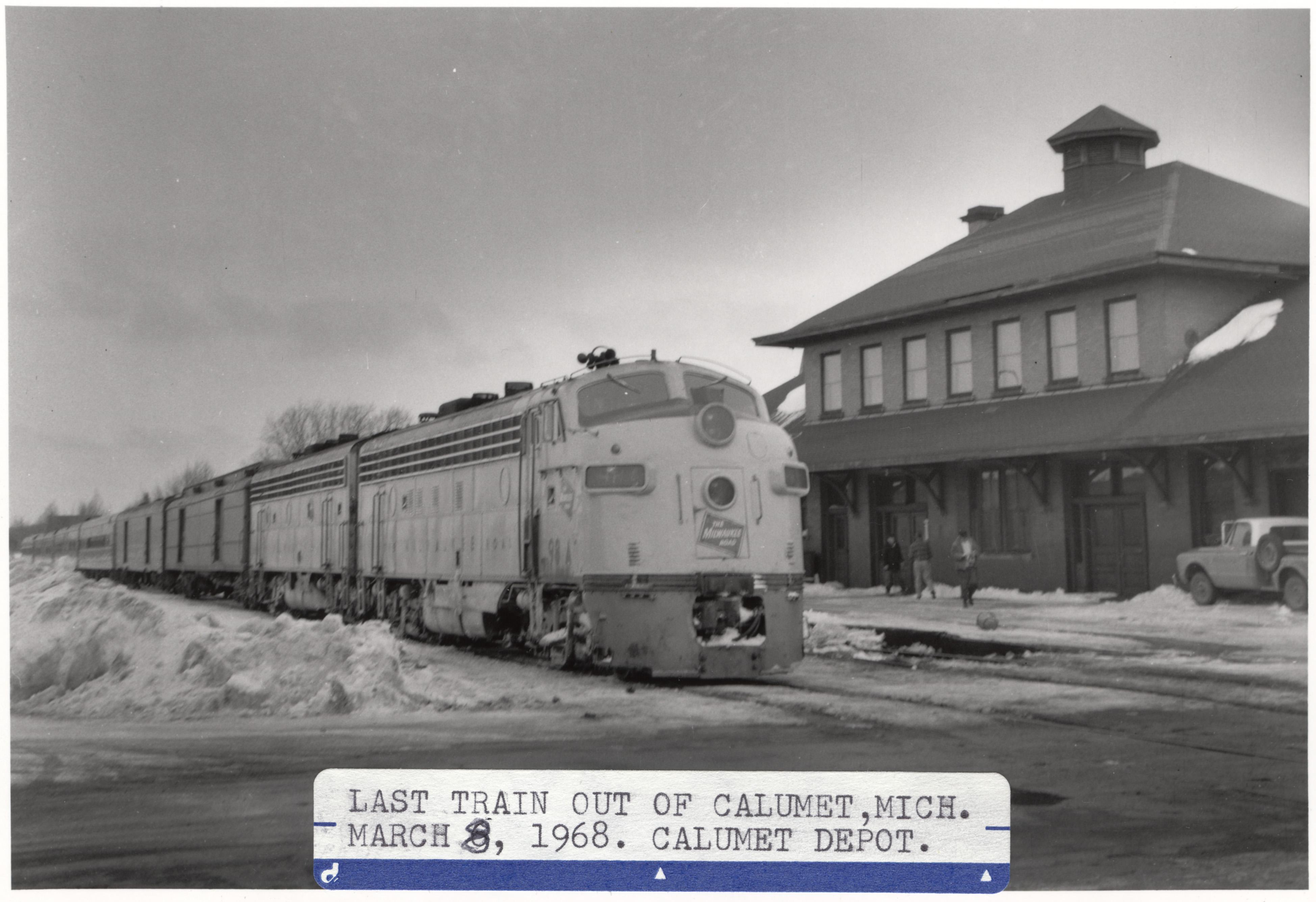 In this black and white image, a train sits in front of a 2-story depot with many windows.
