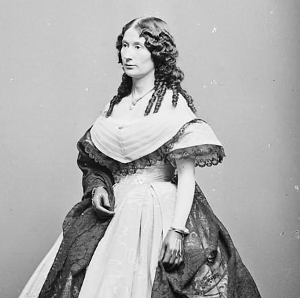Laura Keene standing facing left in dress with large skirt, hair down in ringlets