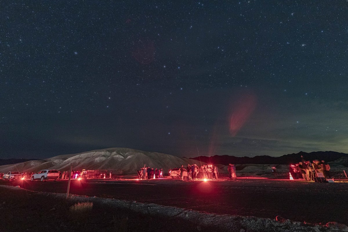 Various individuals use red light at night among many stars and a hilly landscape.