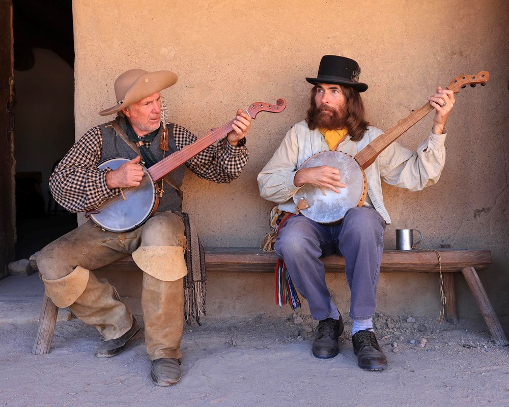 Two musicians sitting on a bench in 1843 period clothing playing banjos