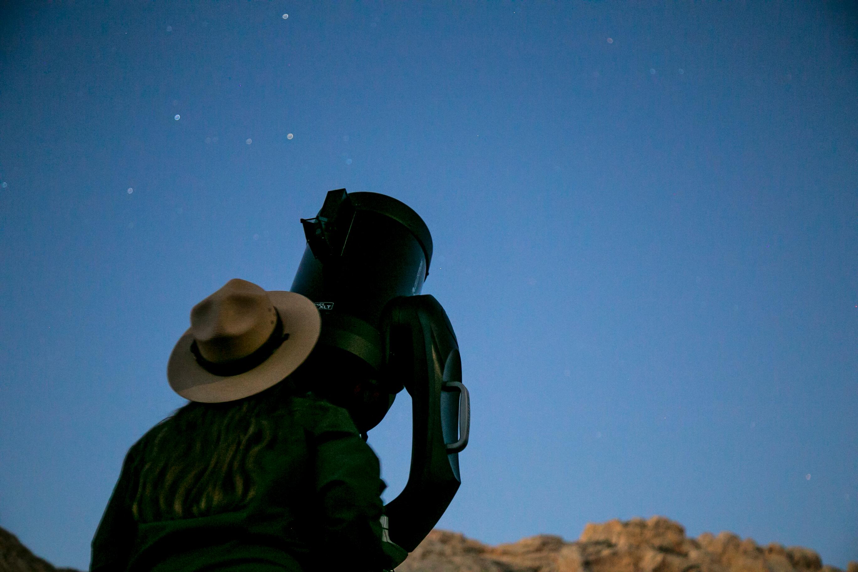 A ranger looks into a telescope as stars begin to come out of a navy blue sky.