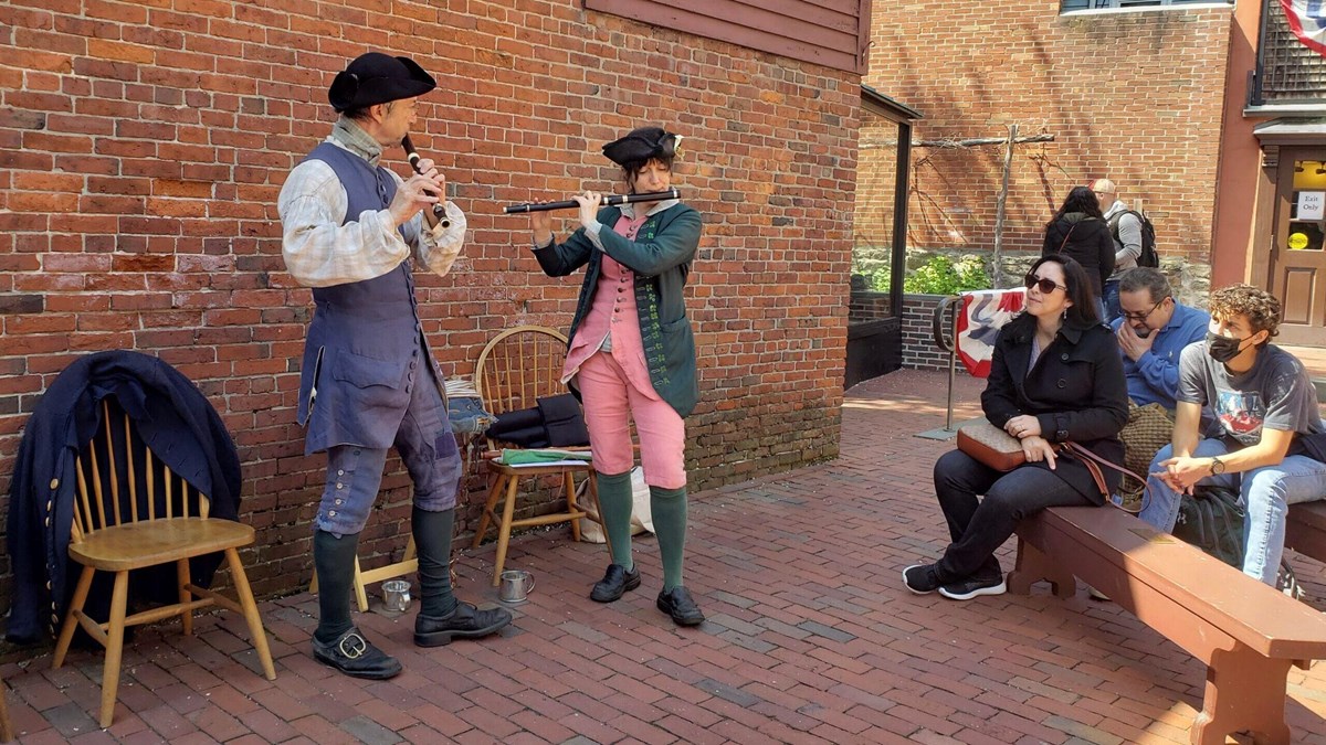 Two people dressed in colonial clothing play historic flutes