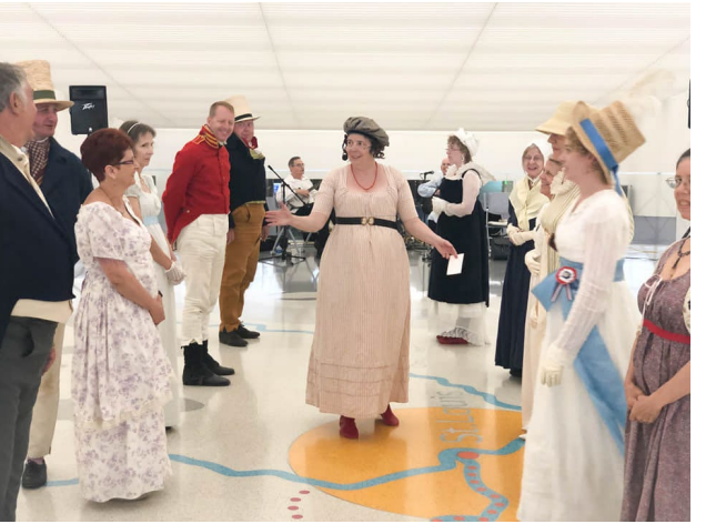 A woman wearing 1800s clothing walks between two rows of other people, also dressed in 1800 clothing