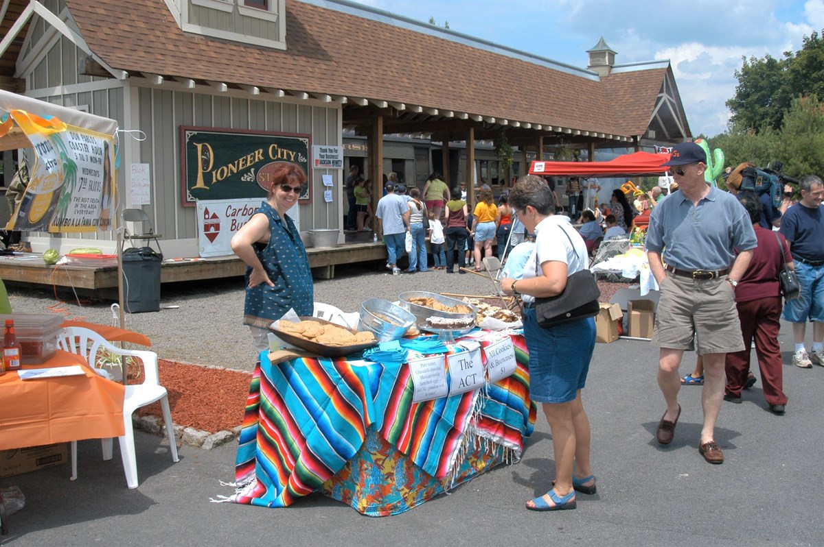 Two people check out a table staffed by a volunteer as part of community event. In the background, a