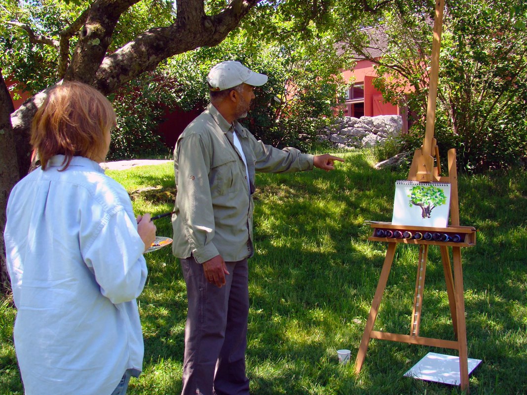A painter is painting in a field on a sunny day while a person looks on and asks questions.