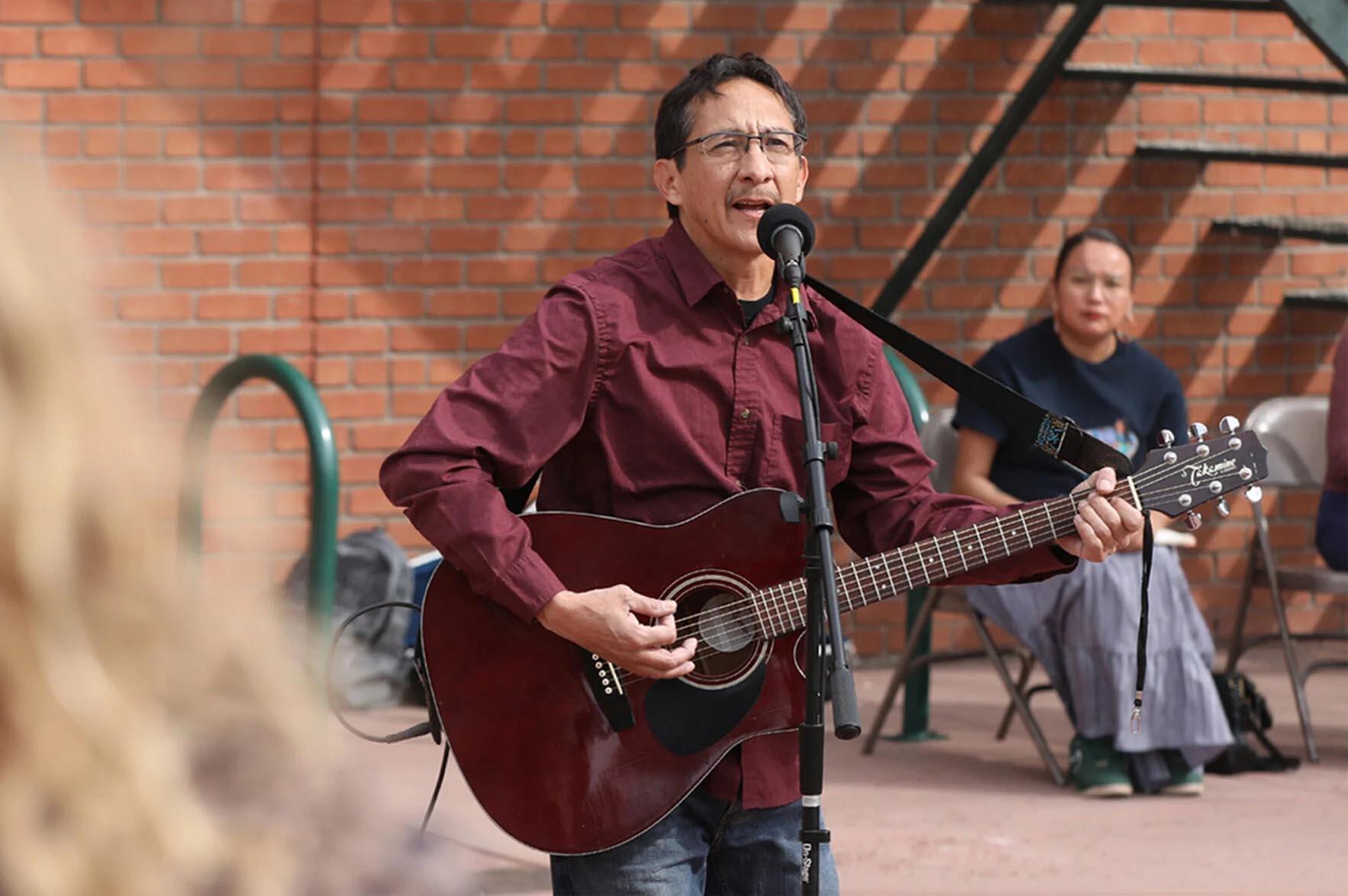 a slender man with black hair, glasses and wearing a maroon shirt, is standing and playing a guitar
