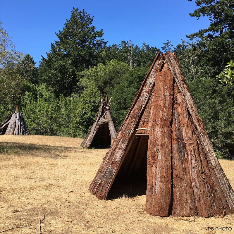 Three ten-foot-tall cone-shaped structures made of redwood-bark sit in a grassy area adjacent to the