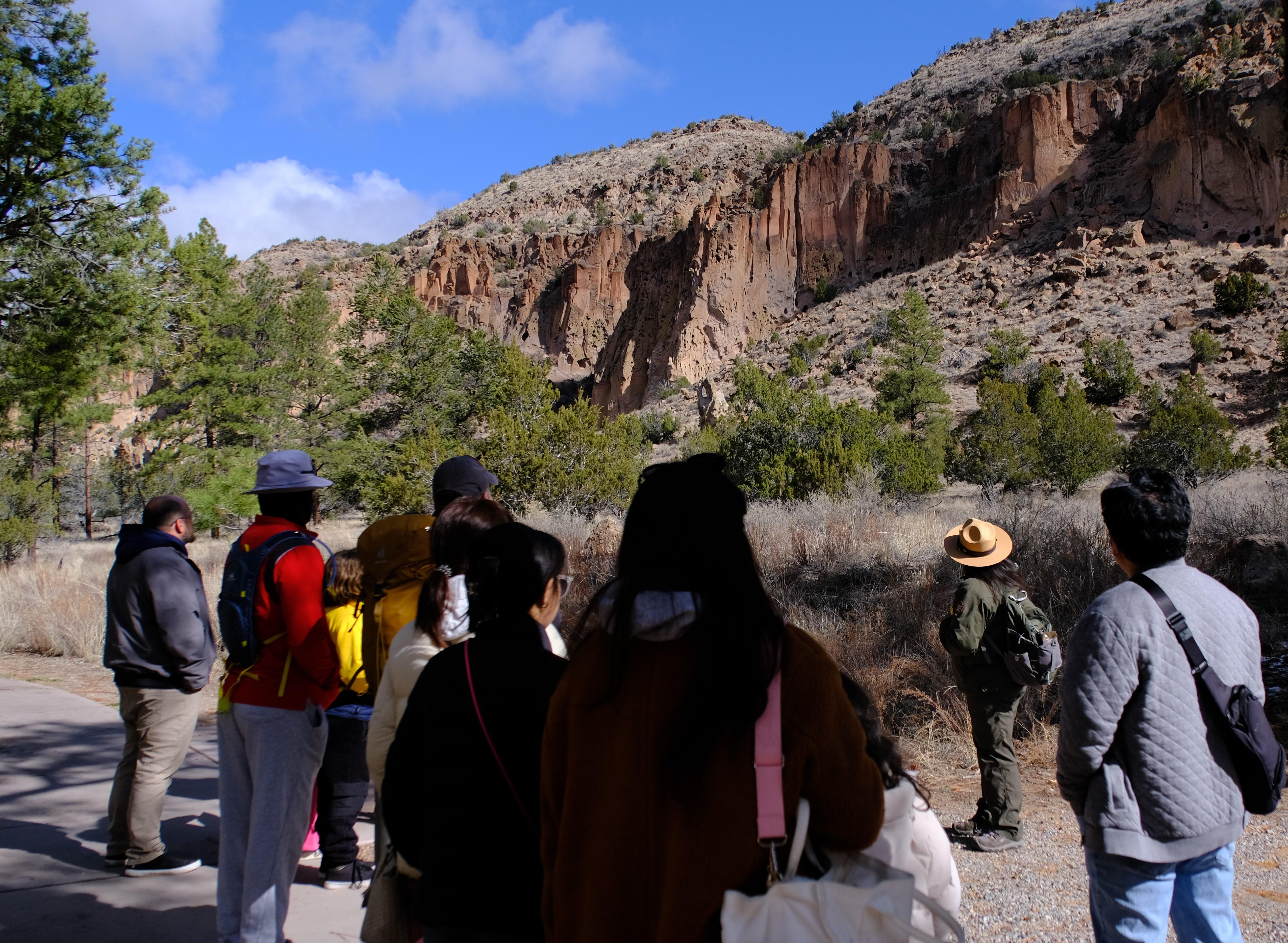 a group of people stand facing a ranger in a green uniform looking up towards a cliff face.