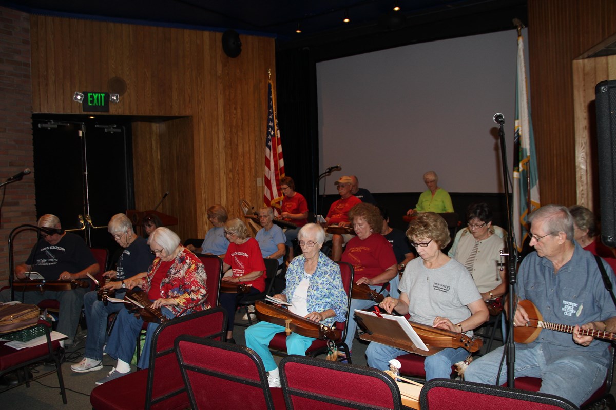 About 15 people, sitting in chairs holding lyre shaped musical instruments in their laps.