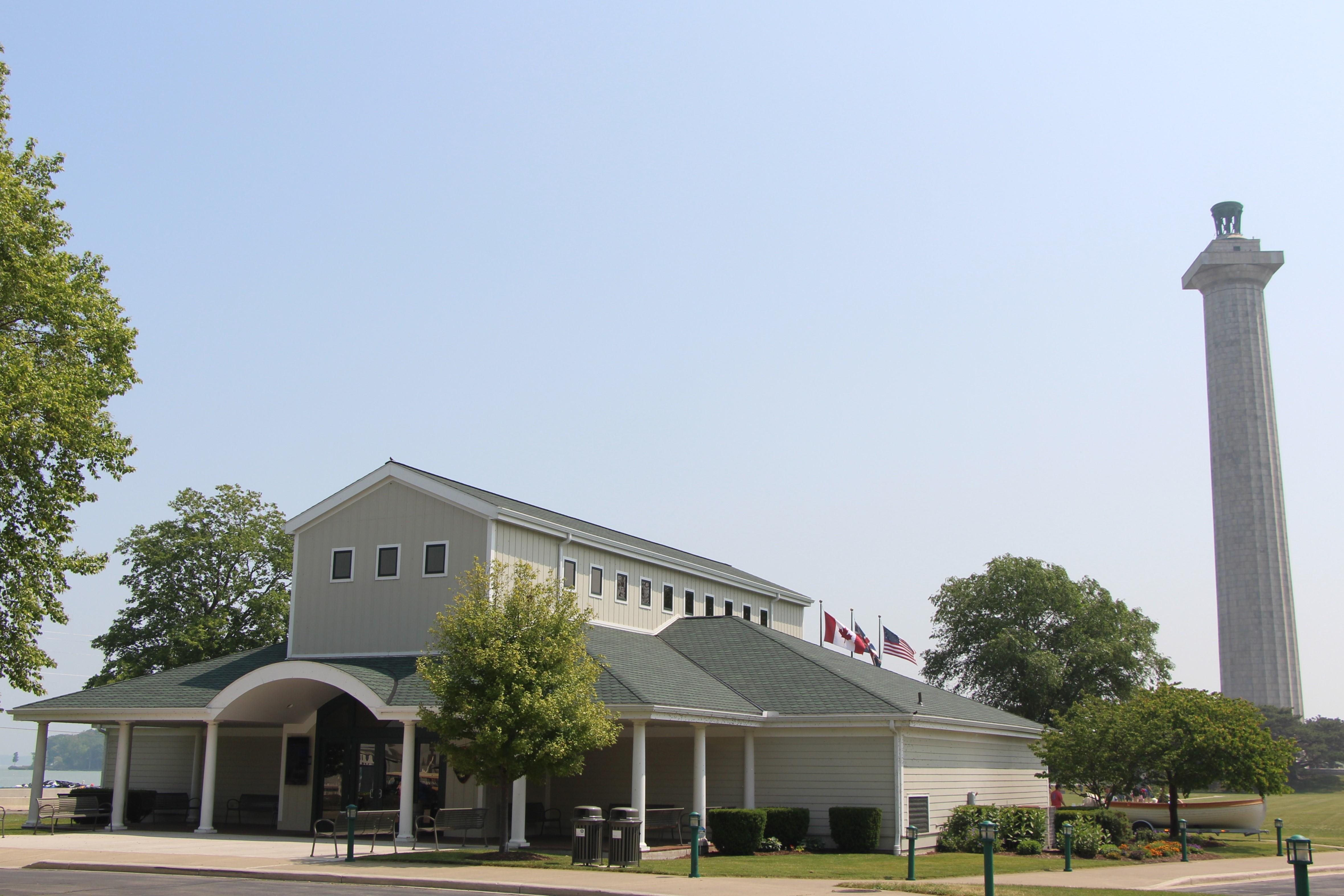 A single-story building with flags and a tall stone monument behind by four trees and a clear sky