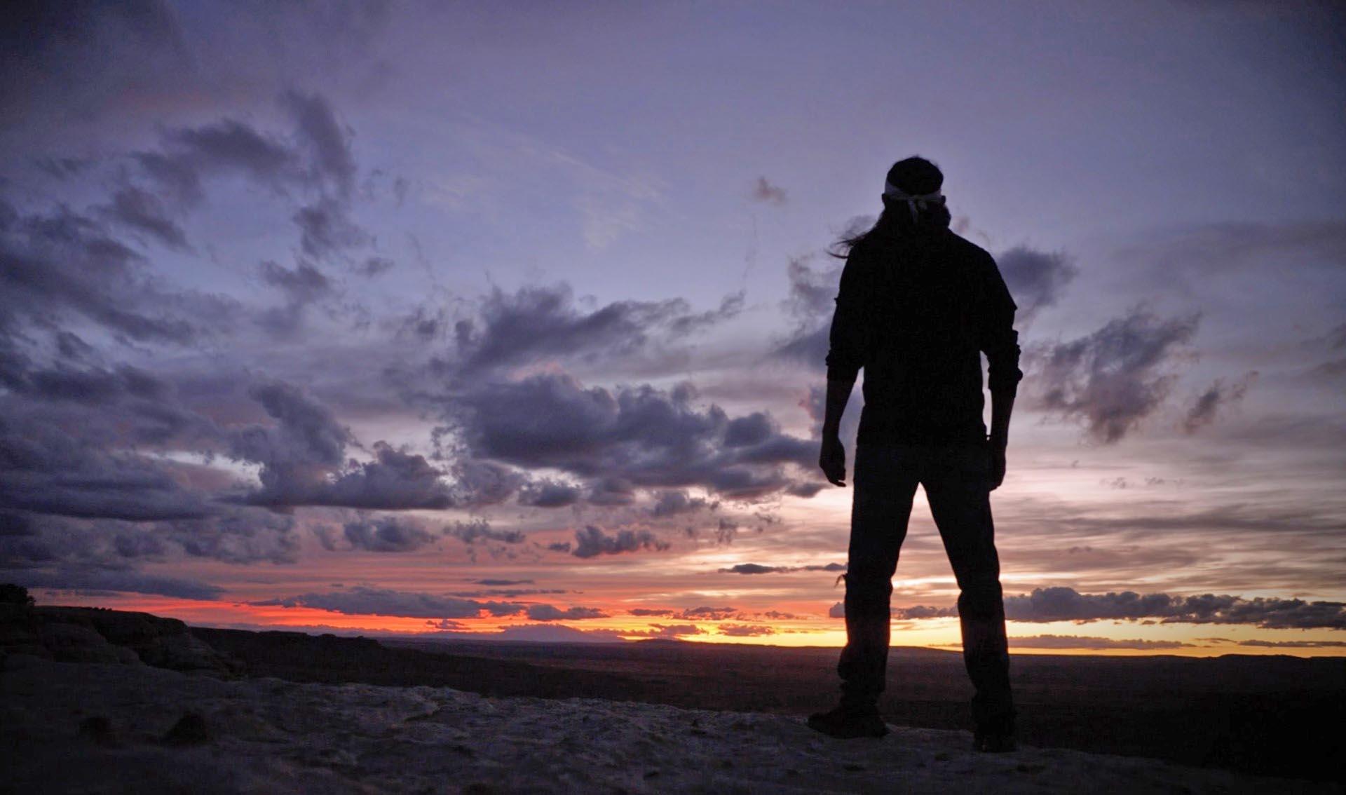 During sunset a silhouetted figure of a person with colorful clouds in the distance.