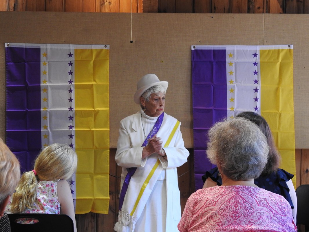 A women in a white dress and hat with with a purple and yellow sash standing in front of a group.