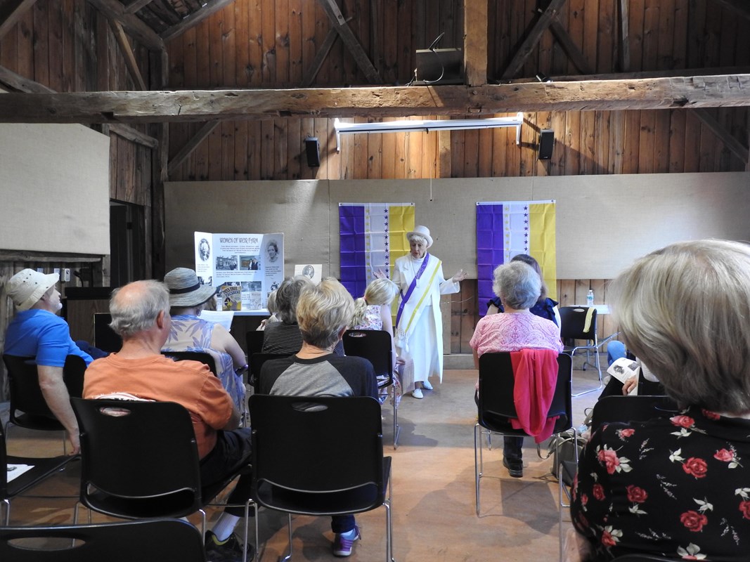 A women in a white dress and hat with with a purple and yellow sash standing in front of a group.