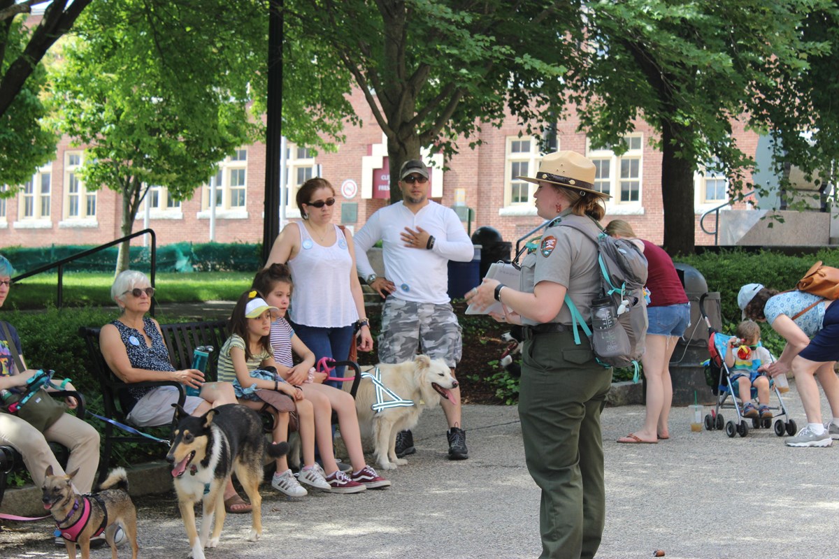 A uniformed park ranger speaks with a group of visitors and their dogs.