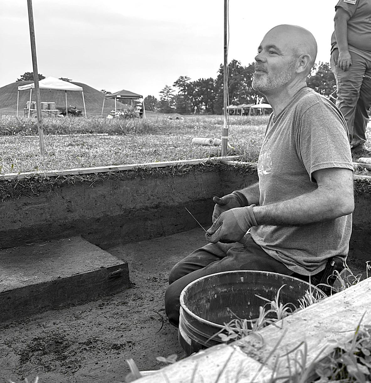 In black and white, a man kneeling in a trench wearing gloves and holding tools for archeology.
