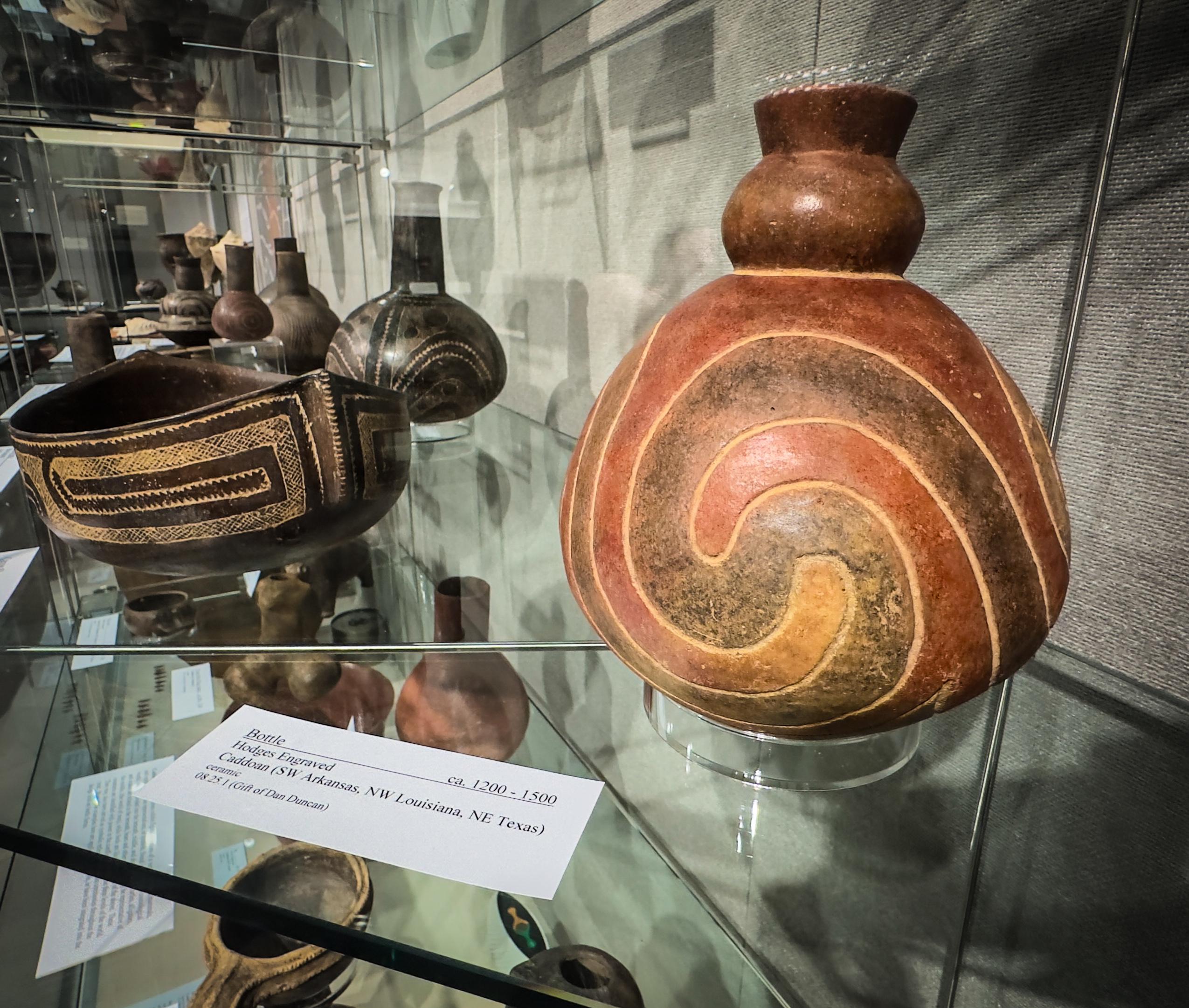 A brown and red clay bottle with a swirl design in a glass display case with a museum label.