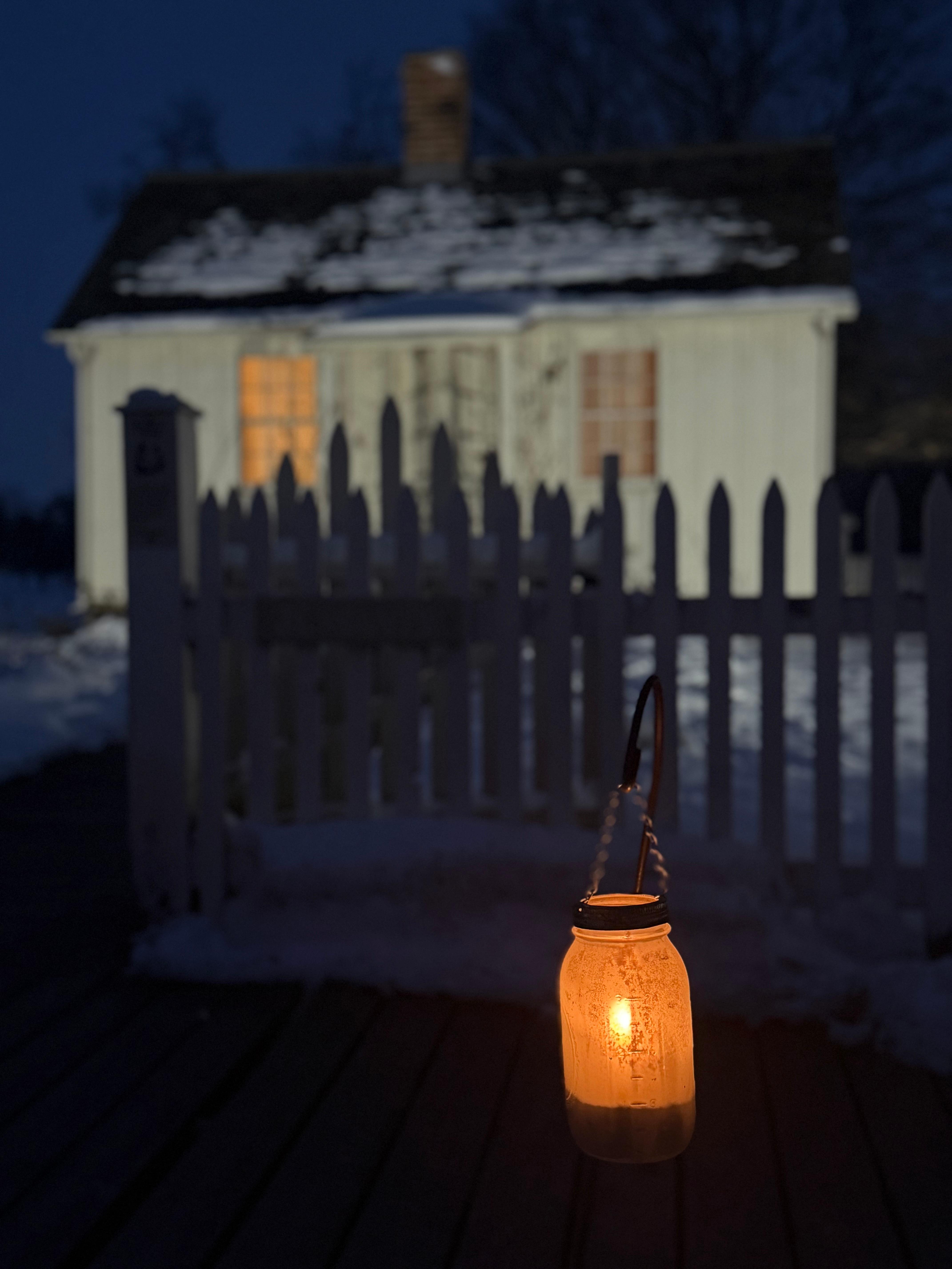 An illuminated cottage and candle light in a glass jar shine bright against a dark night sky.
