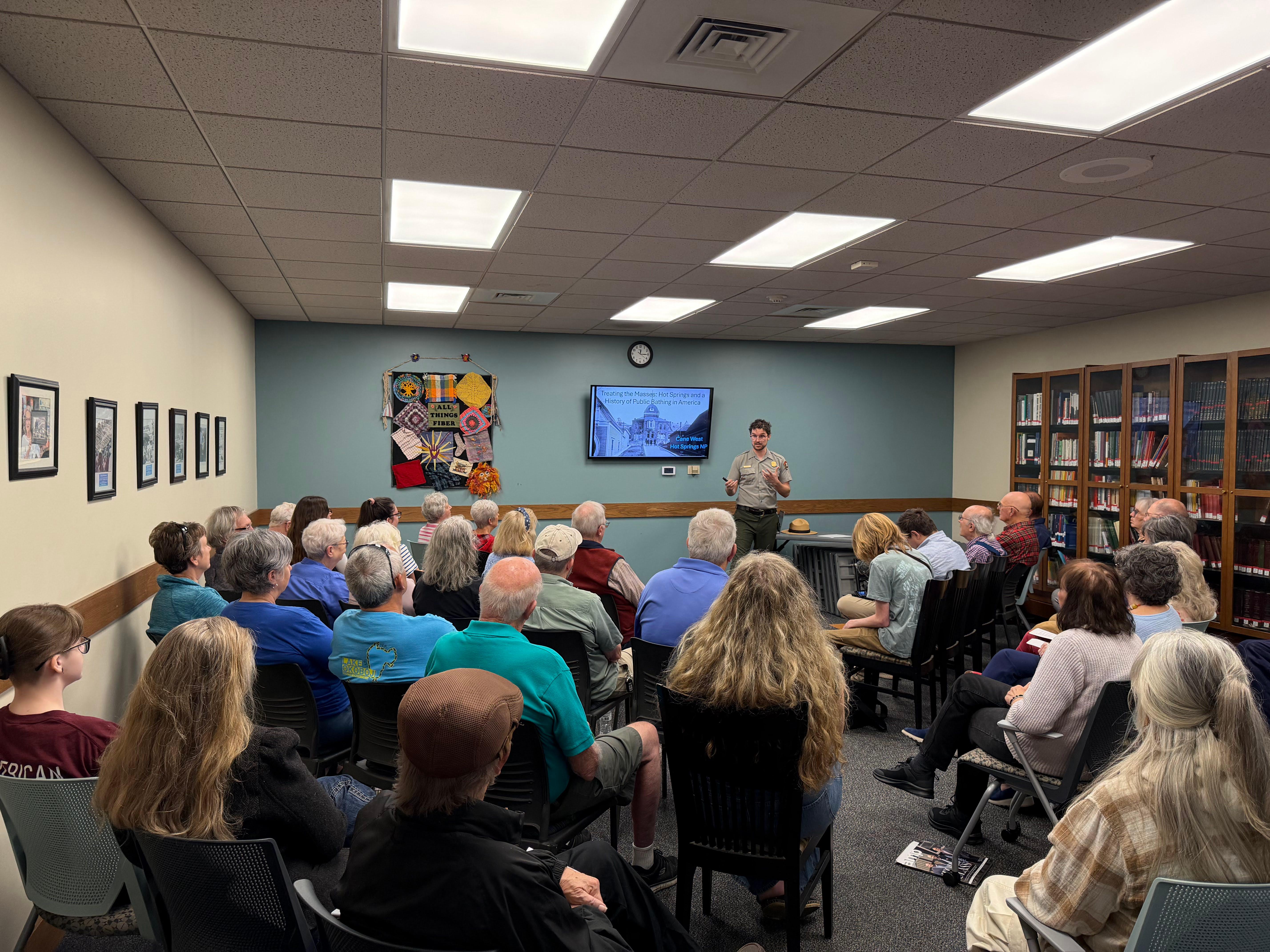 A ranger in a grey shirt speaks to a large crowd of seated people inside of a room.