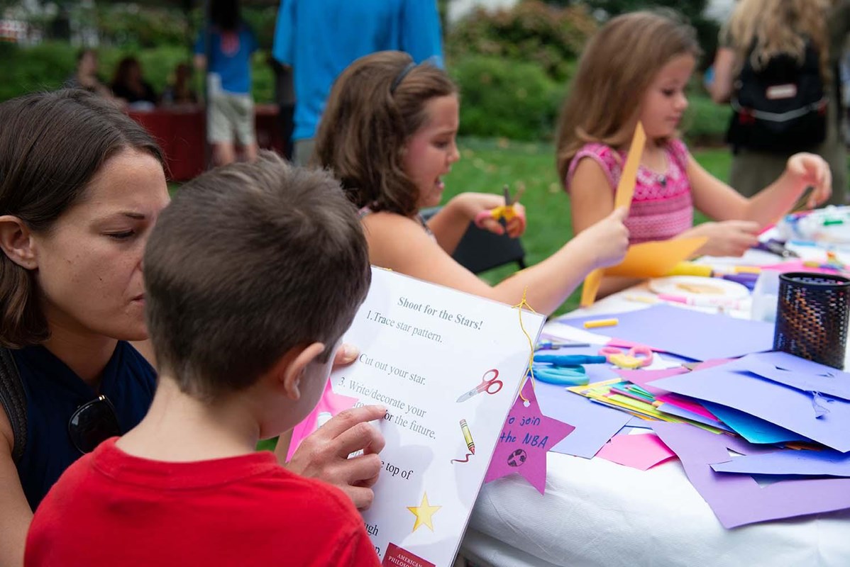 Children make crafts at a table while a woman shows the instructions to a young child.