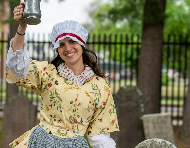 Smiling woman in a colonial American cemetery holding a pewter mug.