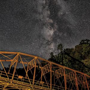 The Milky Way stretches across a star-filled night sky above an illuminated steel truss bridge.