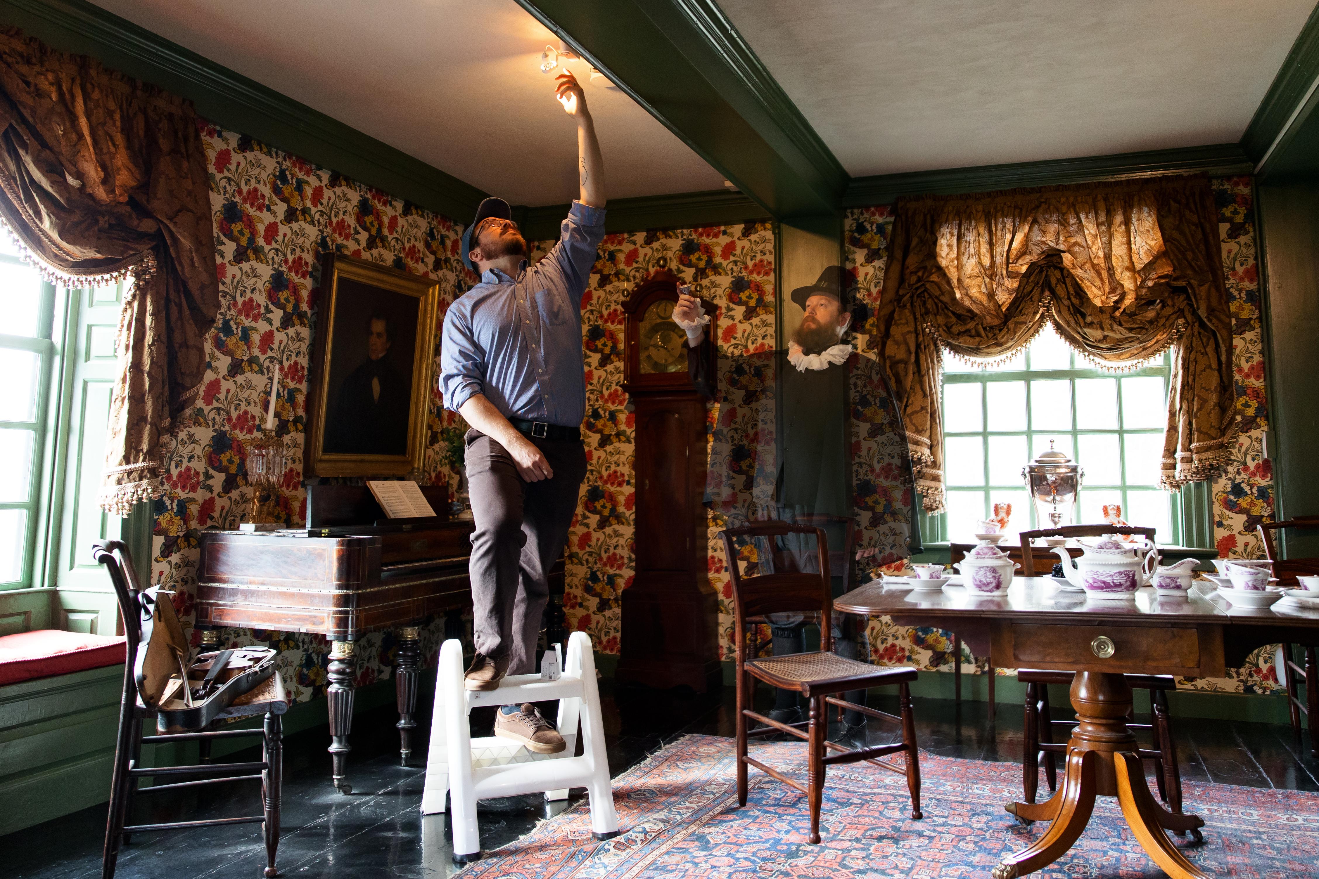 A photo in a historic house shows a man changing a lightbulb with a ghost looking on.