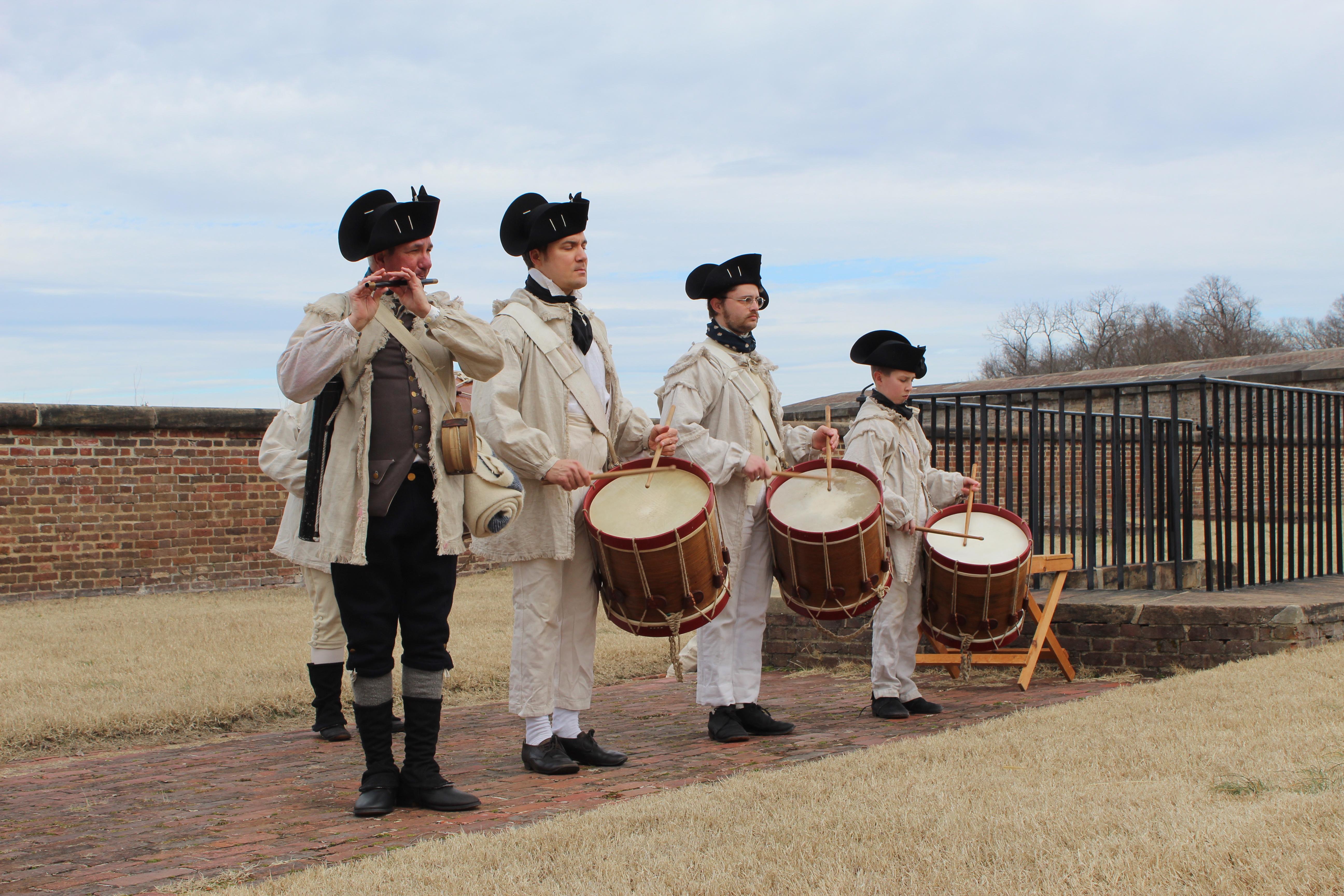 four Fife and Drum Musicians standing in a line with the fort wall in the background