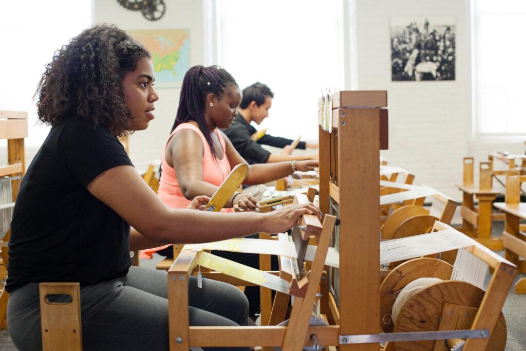 Young people sit at hand looms, weaving their own cloth.