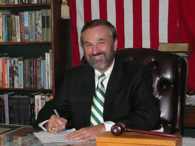Man with dark hair seated at a desk with an American flag behind him