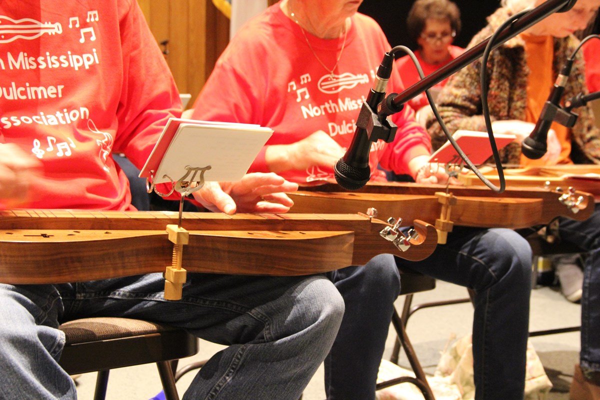 A close up of 3 sitting people in orange shirts. In their laps are wooden lyre shaped instruments.