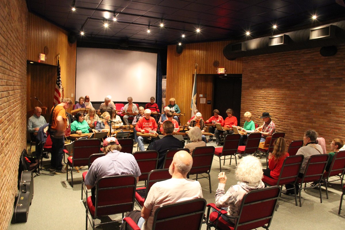 An auditorium with 26 people sitting, playing dulcimers while the audience looks on.