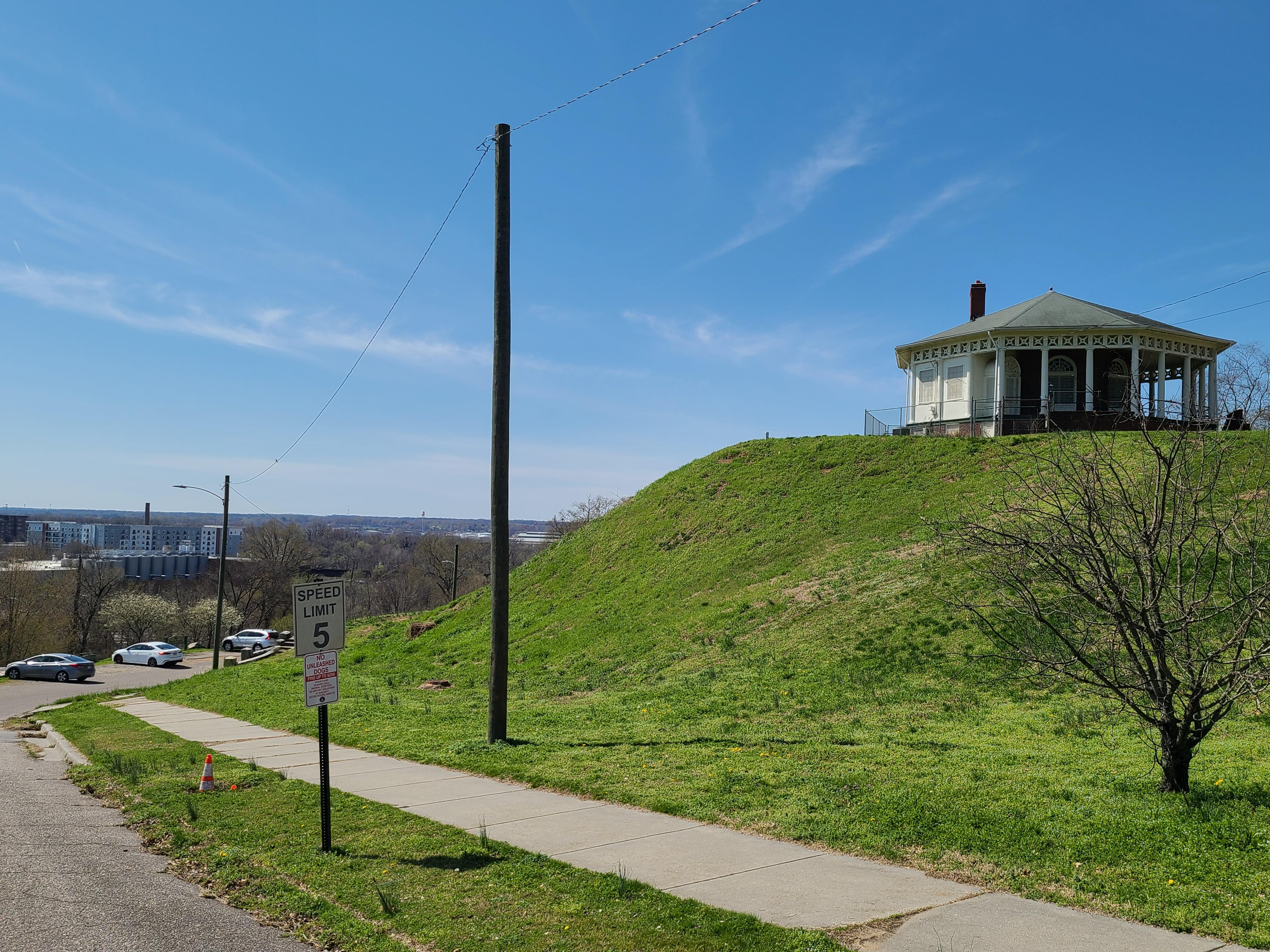 An octagonal red brick building at the edge of a pronounced, grassy hill.