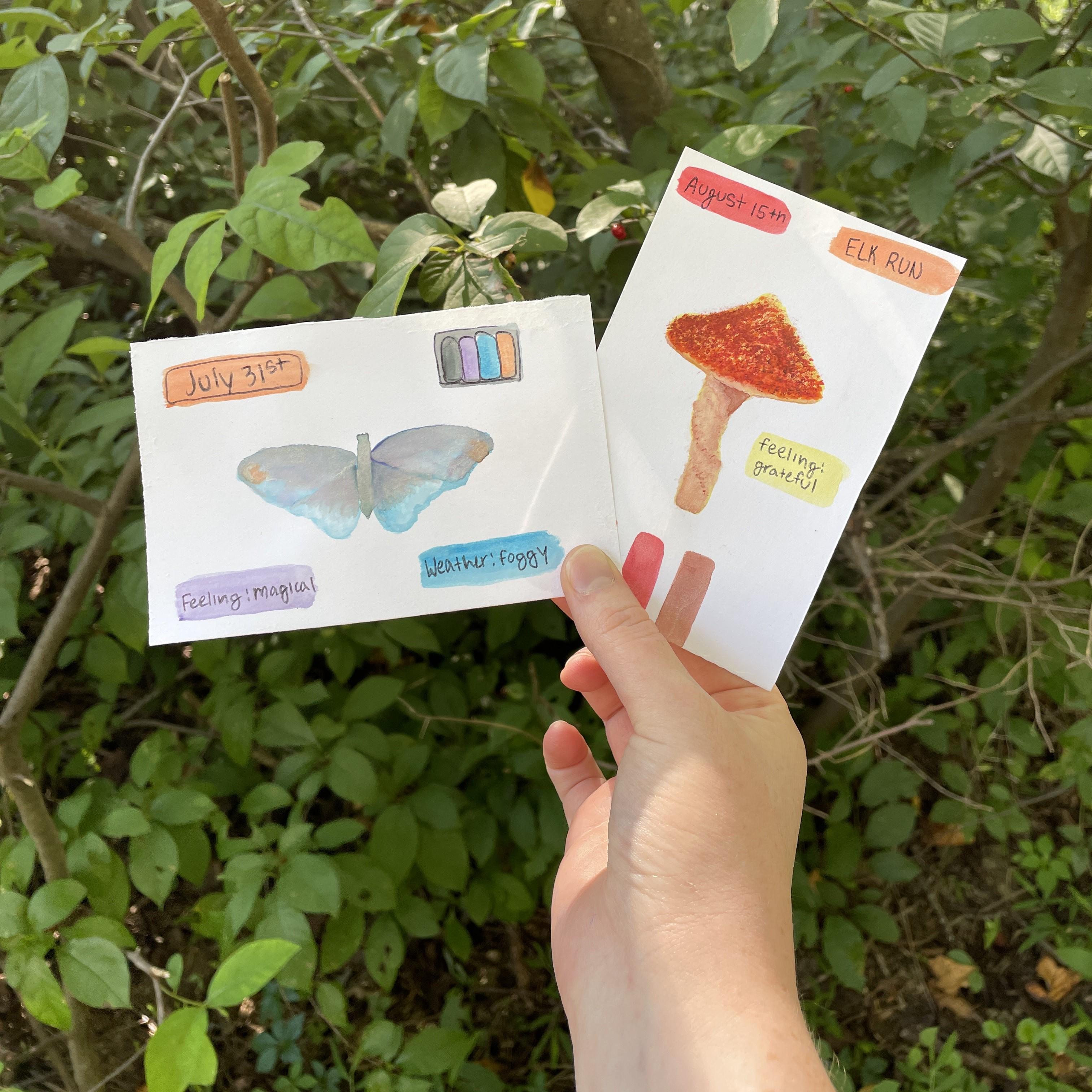 A hand holds up 2 small paintings, a butterfly and a mushroom. Leaves are in the background.