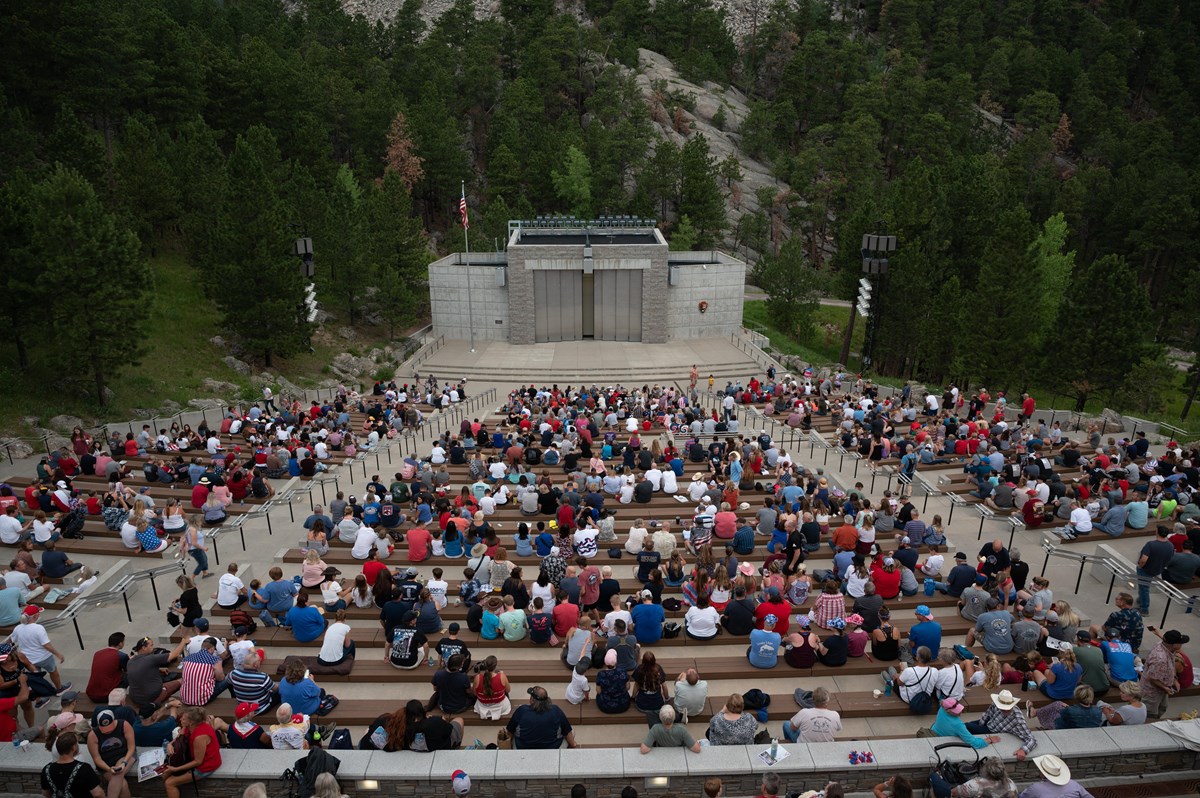 Hundreds of people sitting in a concrete amphitheater with green pine trees on either side.