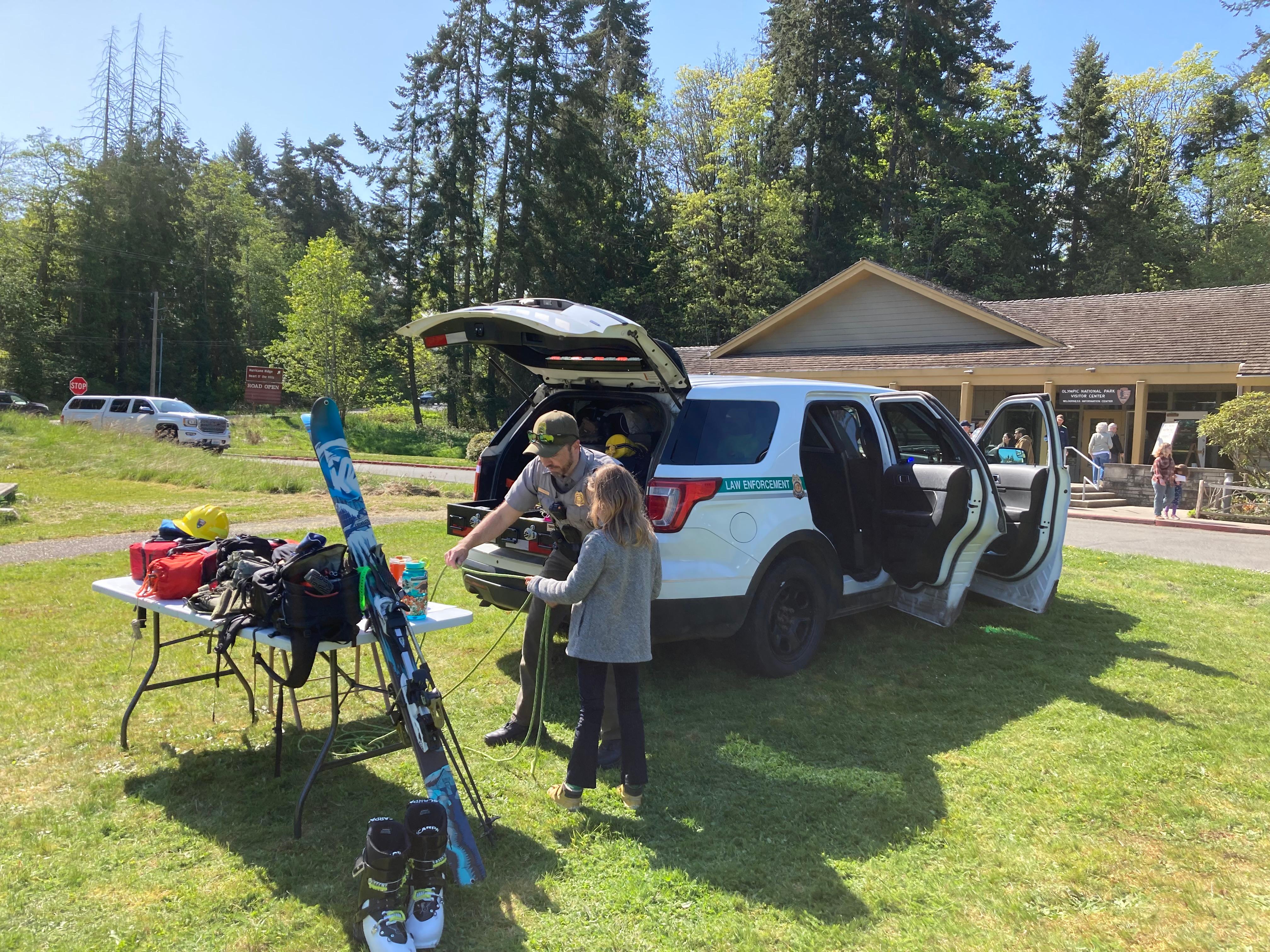 Law Enforcement ranger engages child in front of their vehicle with job tools including skis/ropes.