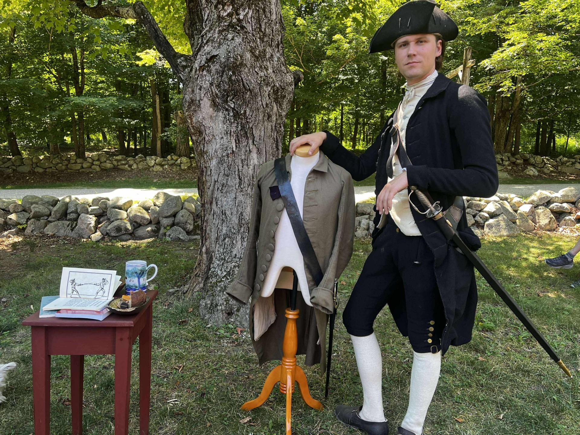 A man in 18th century clothing stands with a sword next to a fencing target