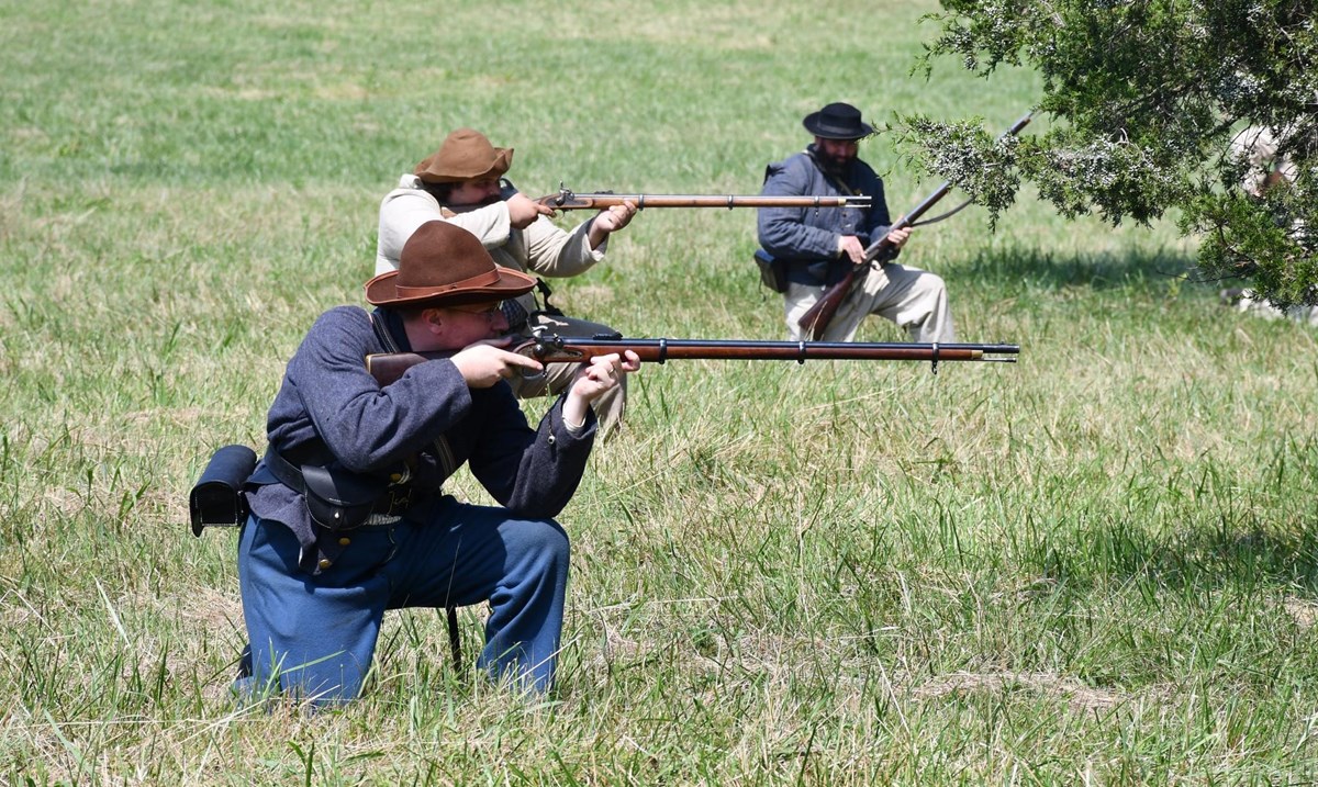 Three white males in Confederate Civil War uniform demonstrate a kneeling position to fire a musket.
