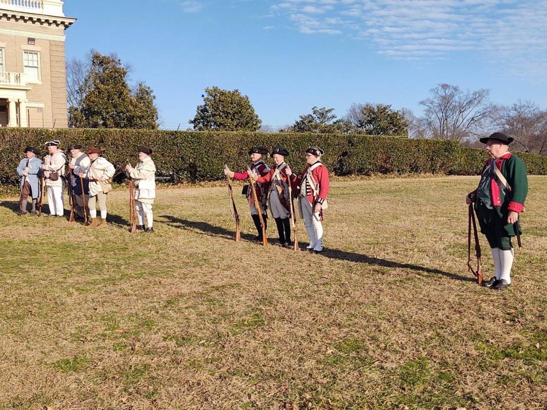 Nine men in Revolutionary War attire stand in a grassy field.
