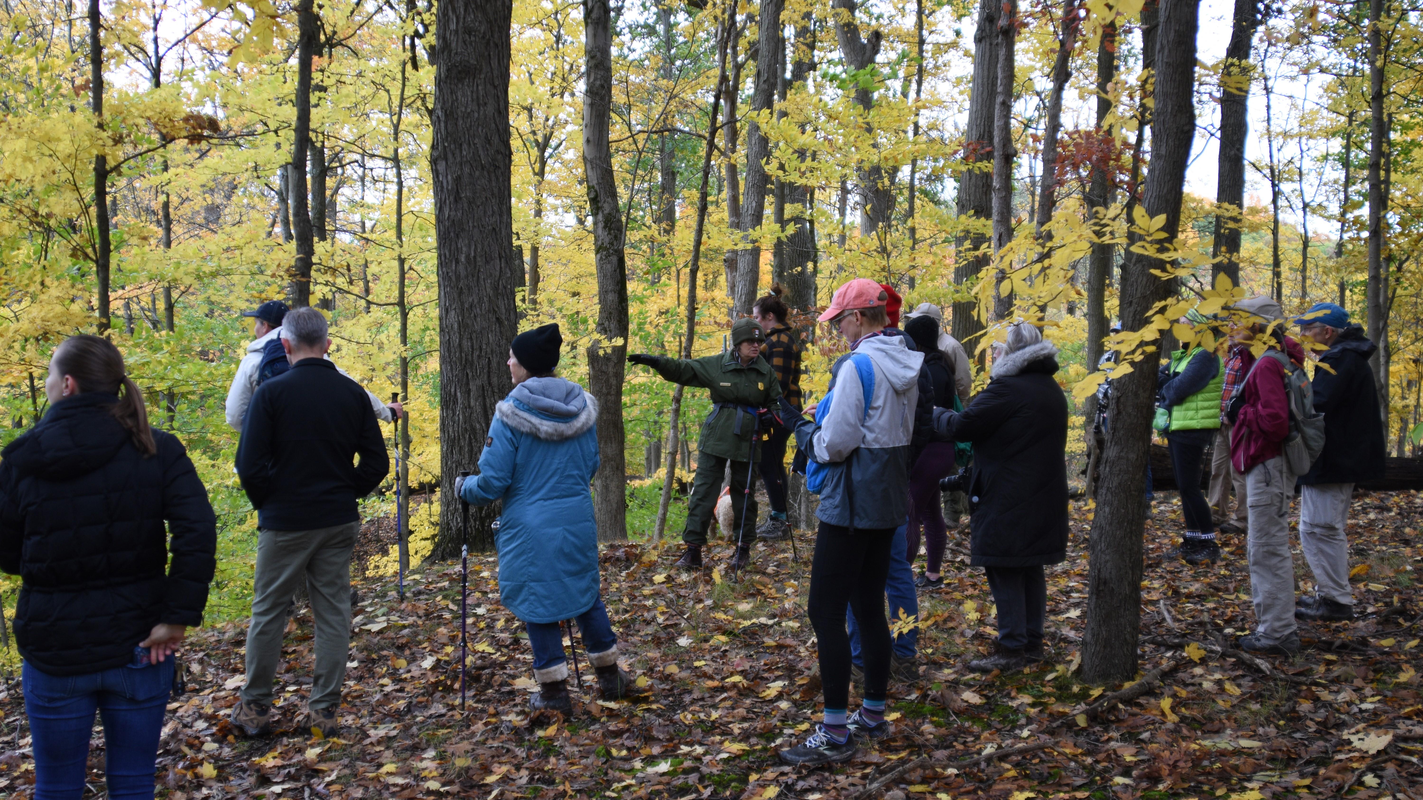 A ranger and a group of 14 hikers stand surrounded by trees with fall foliage.